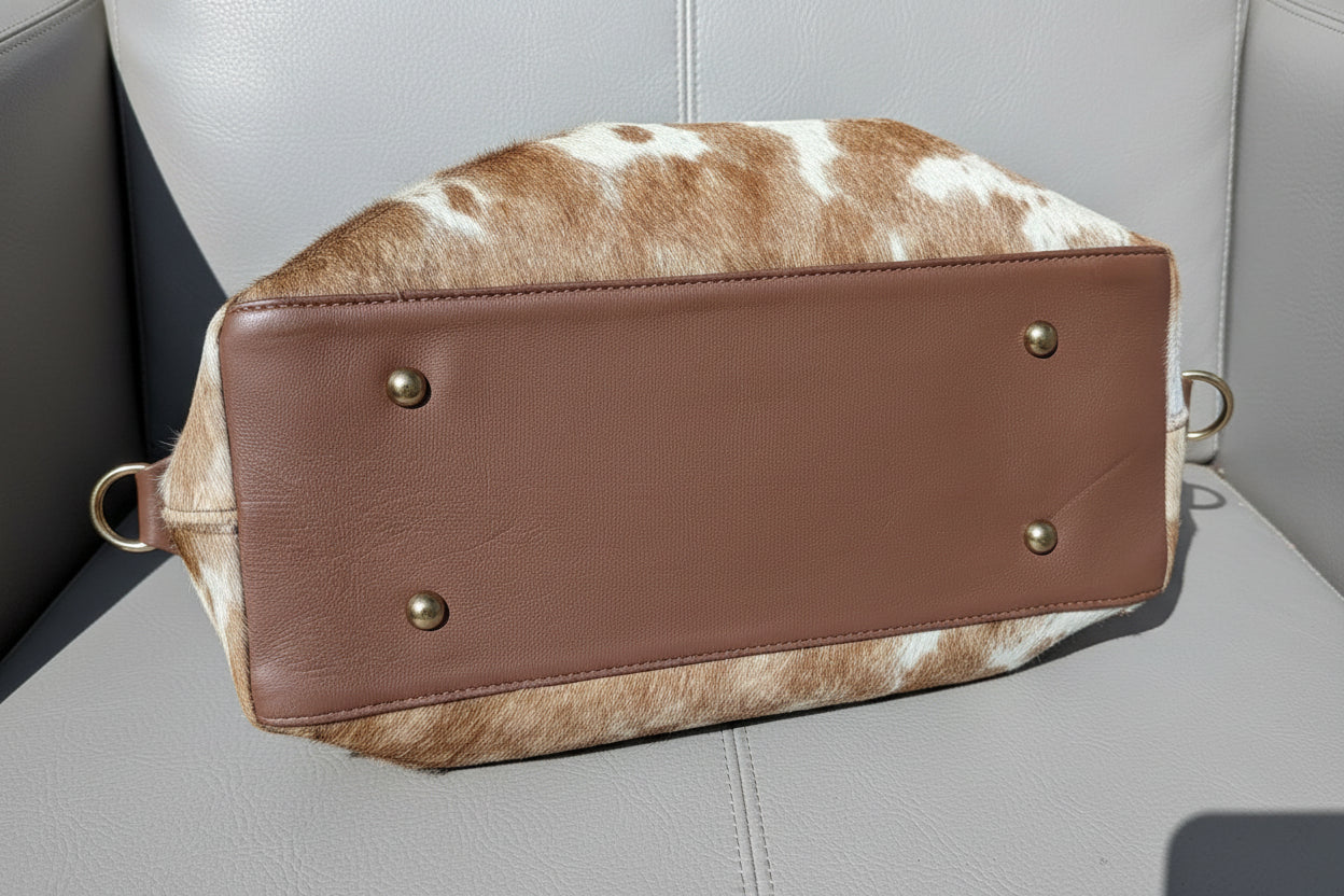 A brown leather bottom panel of a handbag with gold-tone rivets, shown from above on a brown and white cowhide surface over gray paving stones.
