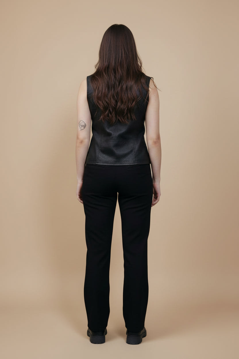 A woman viewed from behind wearing a black sleeveless leather vest and black pants, standing against a solid beige studio background.