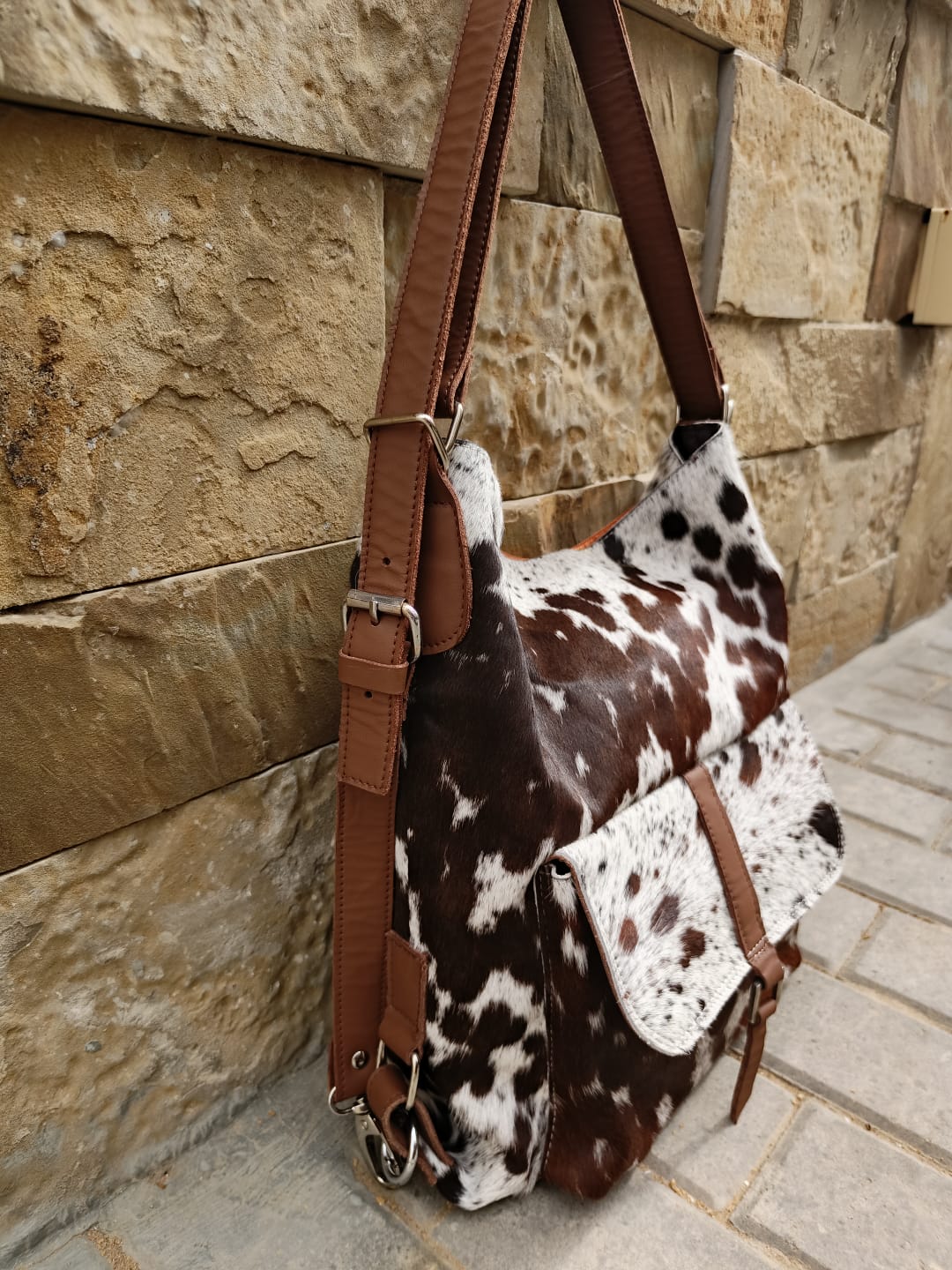 Brown and white cow print bag with brown straps against a stone wall.