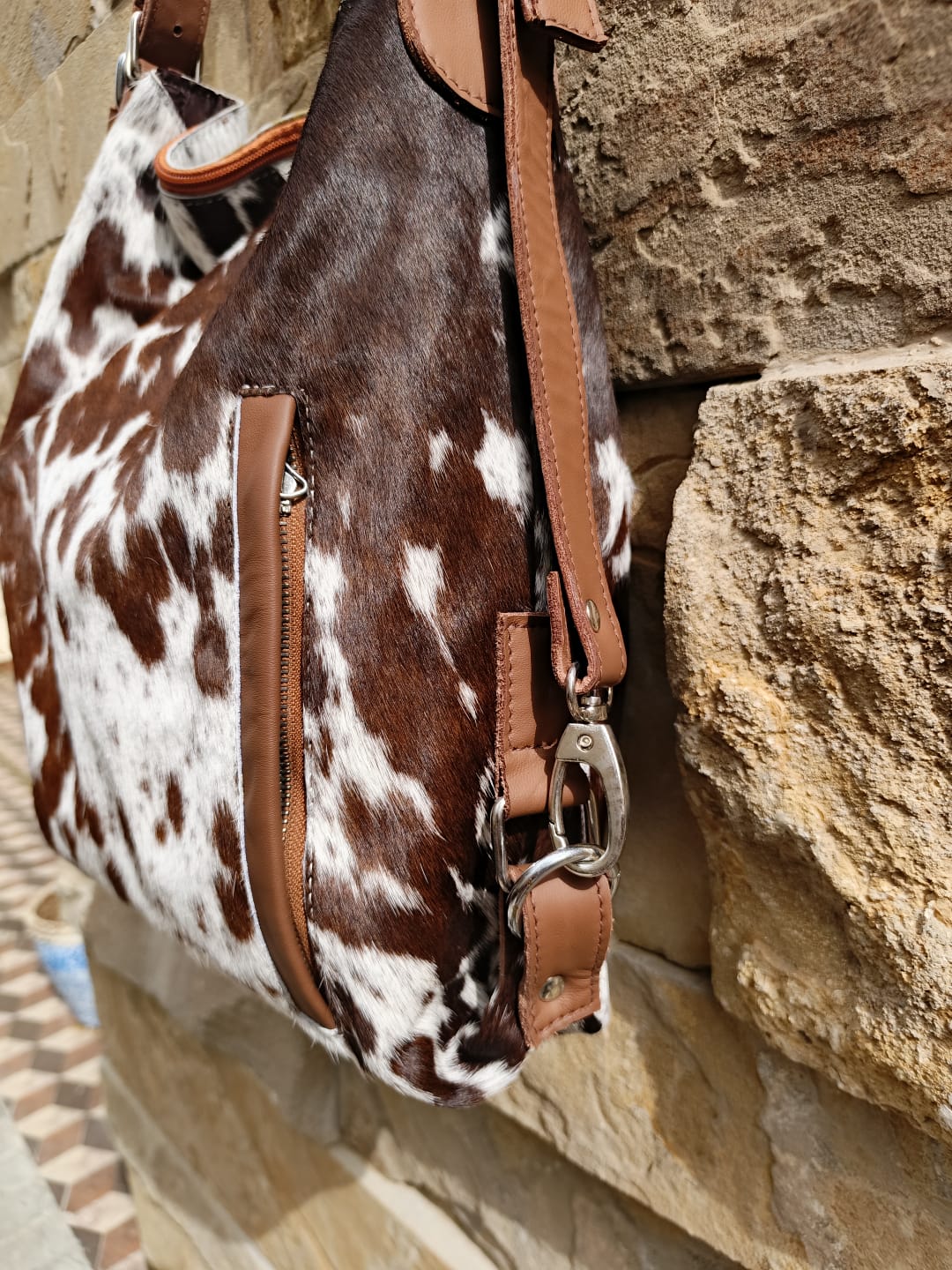 Brown and white cowhide handbag with leather straps against a stone wall.