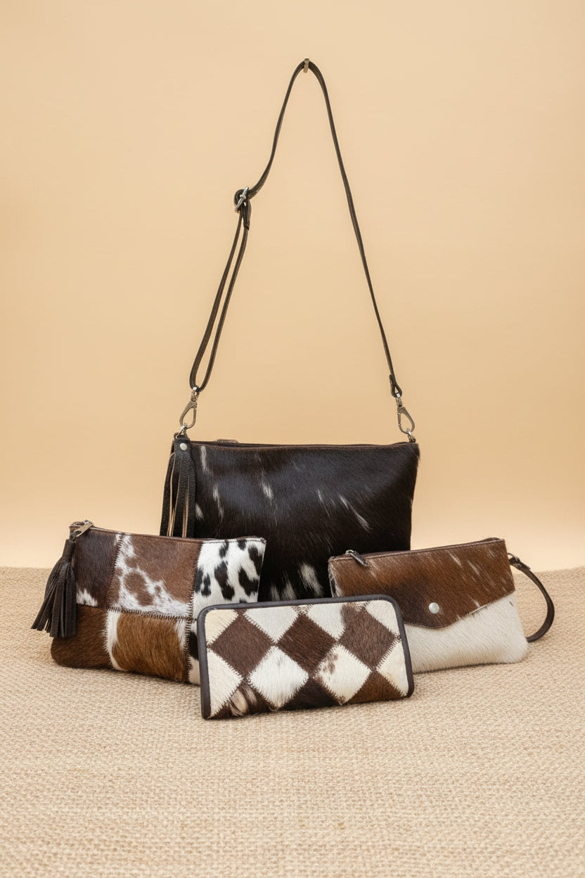 A set of three cowhide-embossed clutch and wallet bags in black, brown, white, and argyle patterns, displayed on a stone patio with plants in the background.