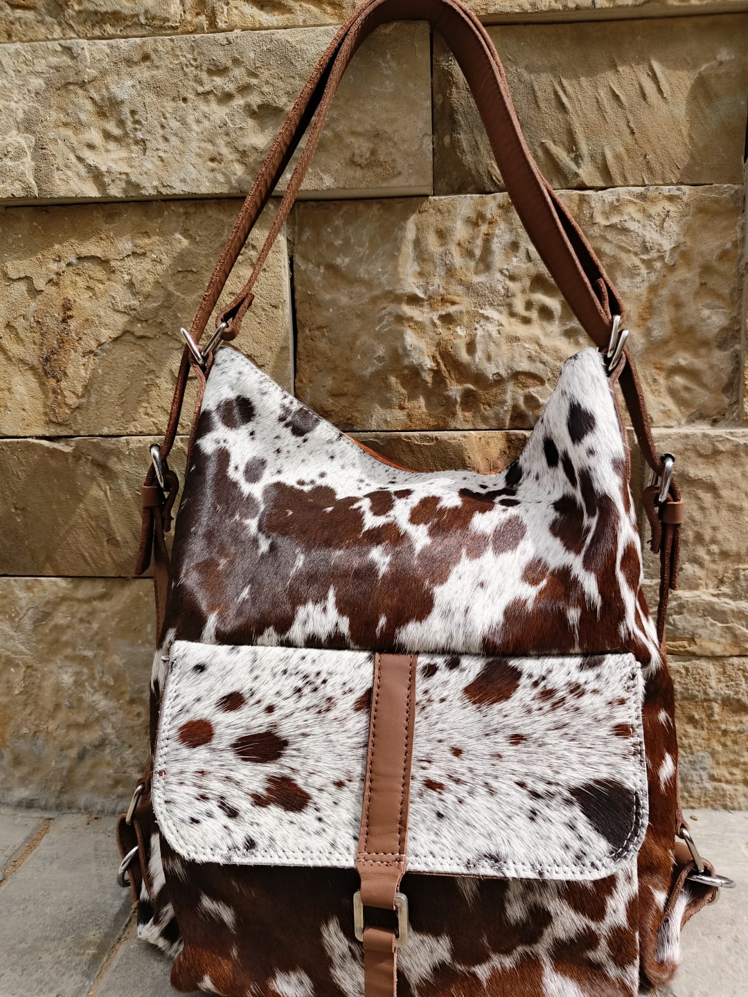 Brown and white cowhide patterned handbag with brown leather straps against a stone wall.