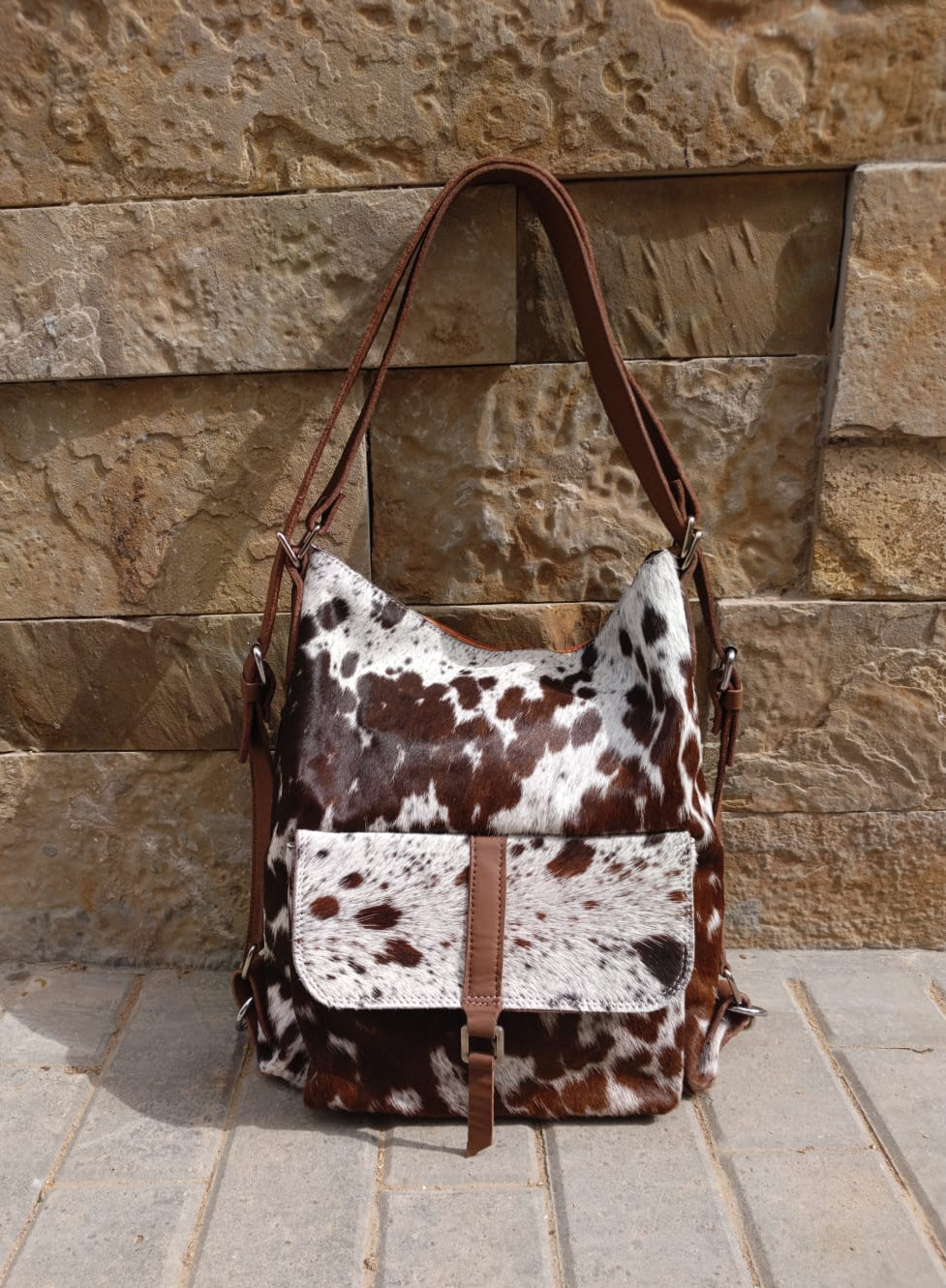 Brown and white cow print bag with brown leather straps against a stone wall.