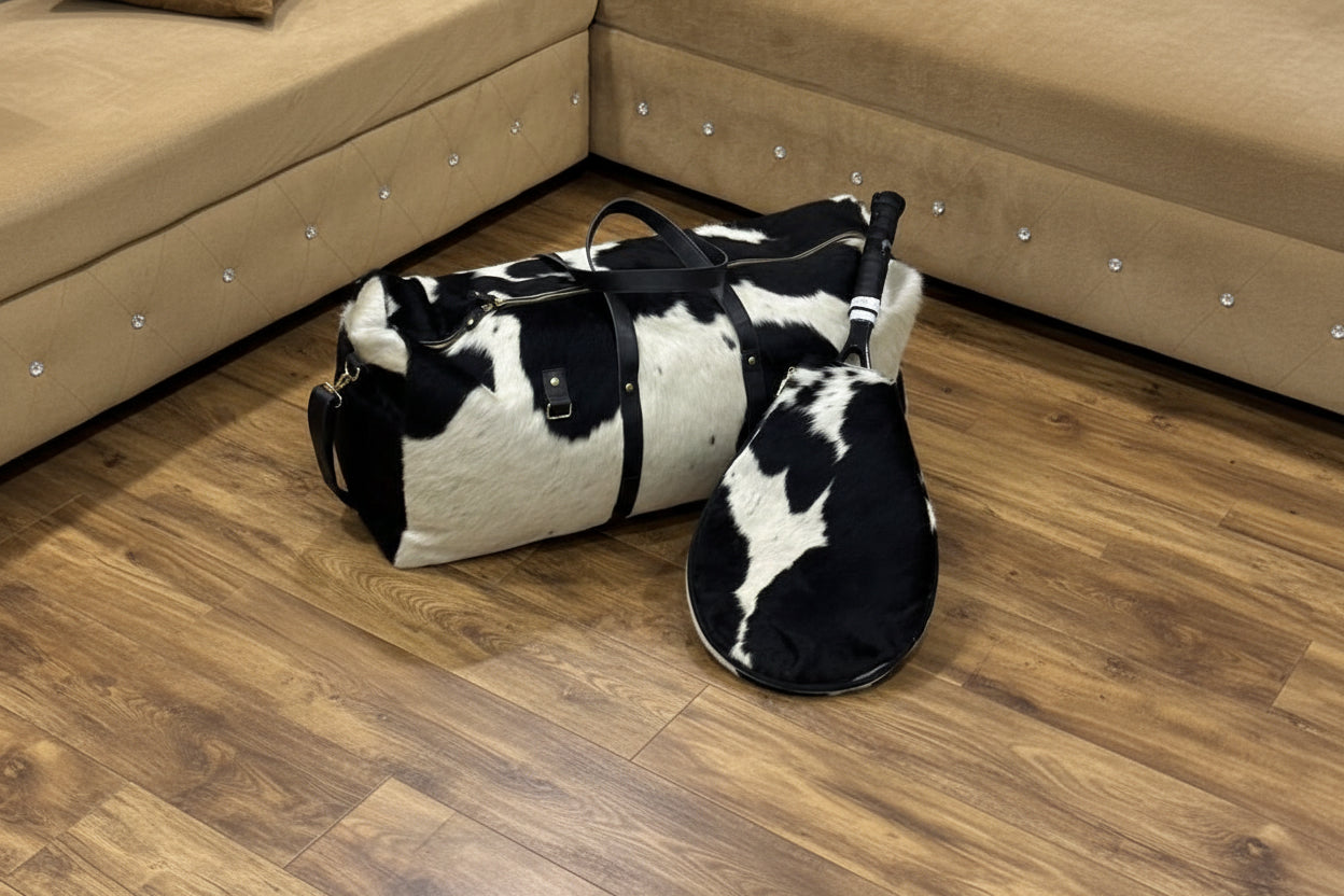 Black and white cowhide duffel bag and slippers on a tiled floor with a beige sectional sofa in the background.