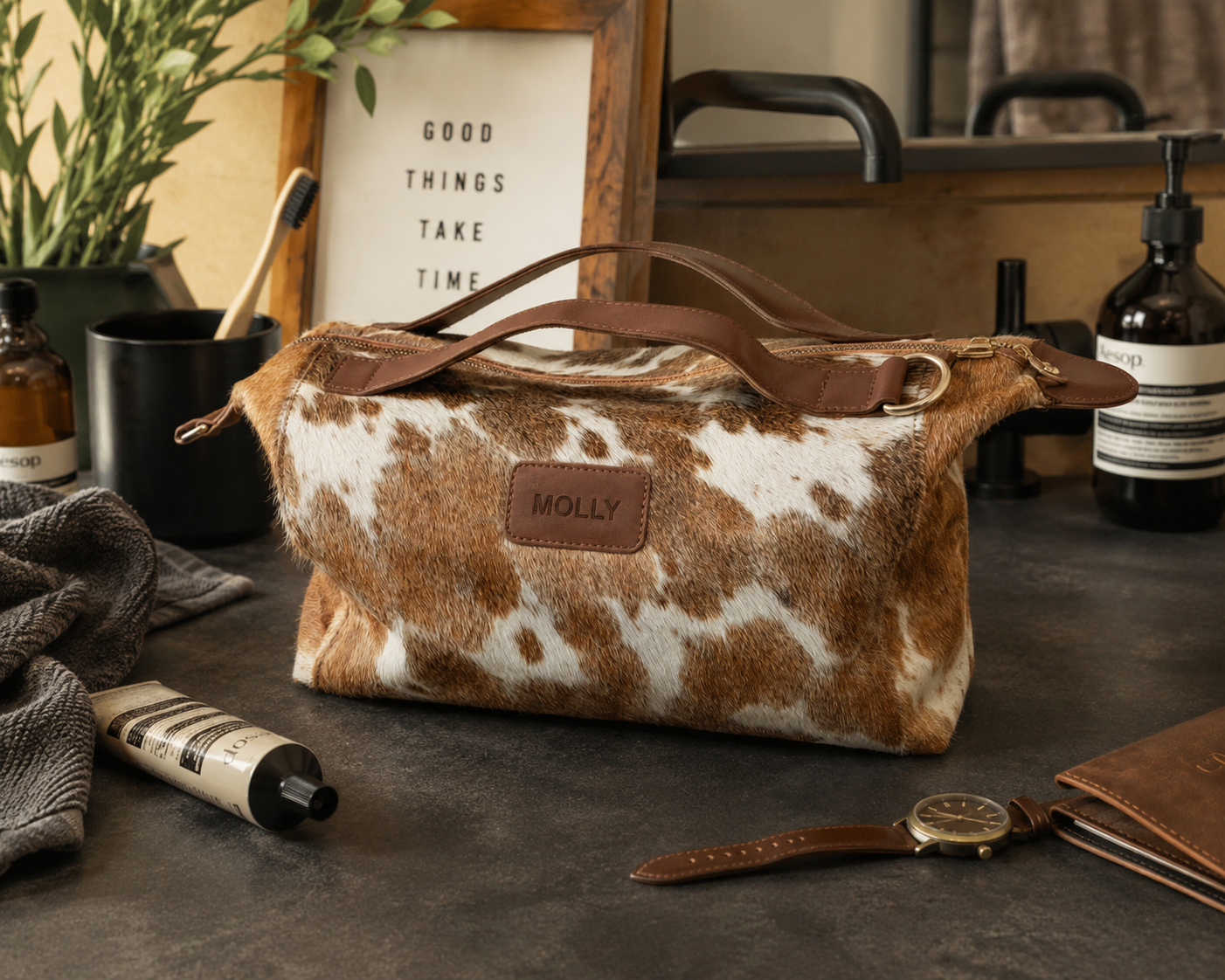 A cowhide leather toiletry bag with brown leather handles and a brown leather patch embossed with 'MOLLY' sits on a dark tabletop in a styled bathroom setting with soap, lotion, and a watch nearby.