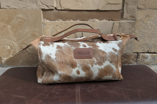 A cowhide duffel bag with brown leather handles and a brown leather brand patch reading 'MOLLY', placed on a stone-tiled floor against a stone wall.
