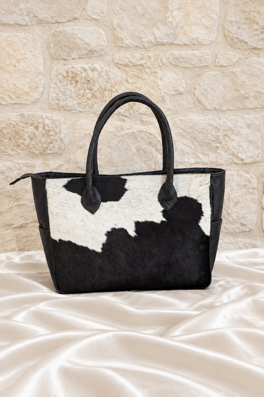 A black and white cowhide tote bag with dark brown leather handles, displayed on a white sating fabric surface against a stone wall